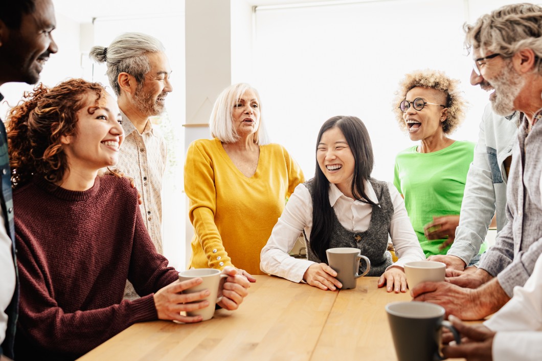 A diverse group of individuals in a business-casual setting sitting around a table drinking coffee and laughing.