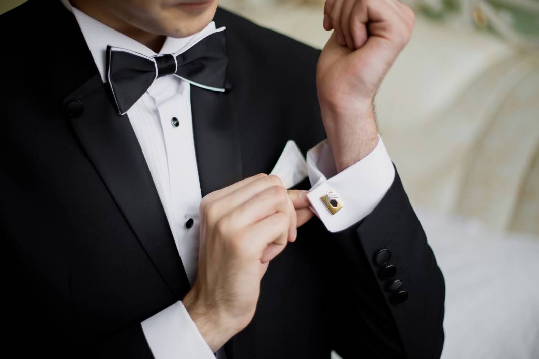 A man in a black tuxedo with a white shirt and black bowtie. He is carefully adjusting a gold-framed cufflink on his sleeve. A man in a black tuxedo with a white shirt and black bowtie. He is carefully adjusting a gold-framed cufflink on his sleeve.