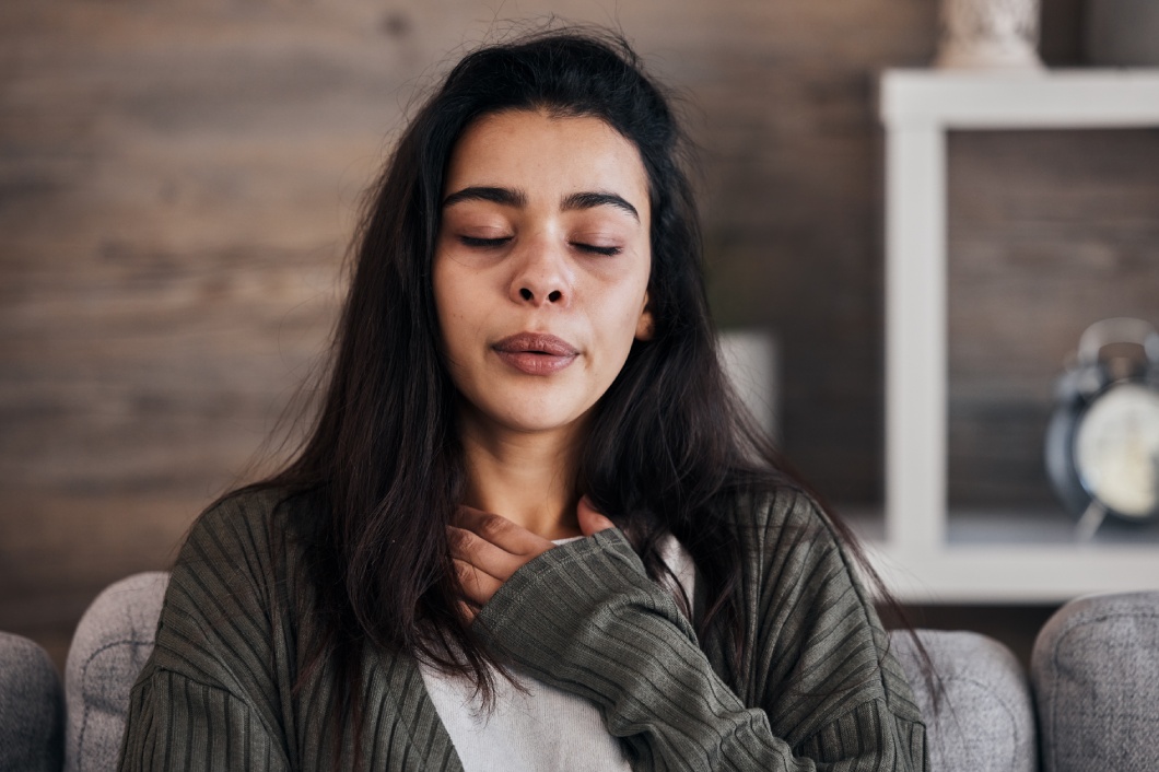 A young woman in a sweater holds her hand to her chest while closing her eyes and breathing through her mouth.